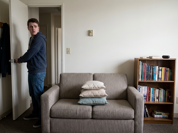 Mladý muž s úzkostným výrazem kontroluje zámek dveří, zatímco v pozadí je vidět dokonale uspořádaný gauč s polštáři a knihy seřazené podle výšky.