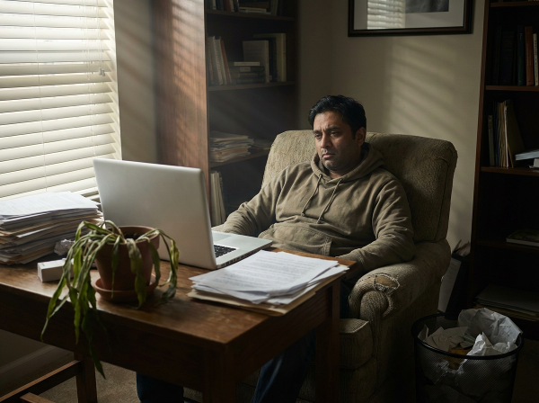 एक 43 वर्षीय पुरुष अपने घर के कार्यालय में अकेले बैठा है, उसके चेहरे पर थकान और उदासी है। वह लैपटॉप स्क्रीन को देख रहा है, जबकि उसके आसपास का वातावरण उसकी भावनात्मक दूरी और तनाव को दर्शाता है।
