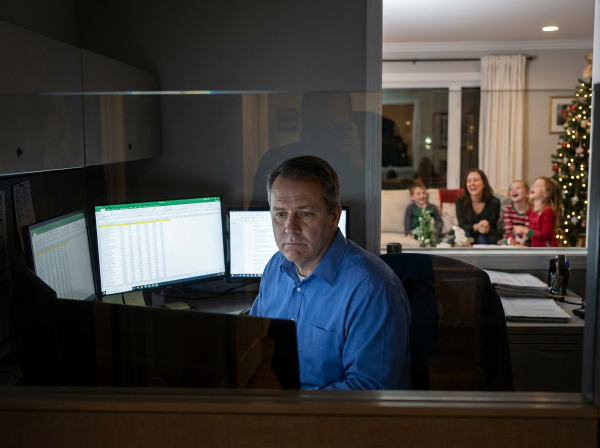 A man in an office looks at a spreadsheet, appearing detached, while a faint reflection shows his family in the background, symbolizing emotional distance.