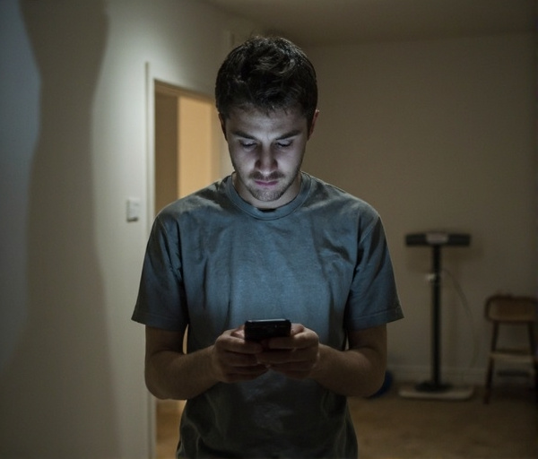 Un giovane uomo siede in una stanza con lo smartphone che mostra grafici di calorie e passi; sullo sfondo una bilancia da cucina e un percorso immaginario di passi, simboli di una routine rigida che occupa la mente e limita la vita sociale.