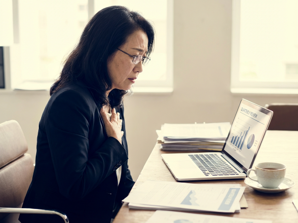 57歳の女性が、プレゼンテーション前の不安を感じながらオフィスで仕事をしている様子。胸に手を当て、緊張した表情を浮かべていますが、窓からの柔らかい光が希望を感じさせます。