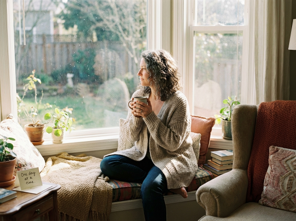 47歳の女性が、穏やかな朝の光が差し込む部屋で窓の外を見つめ、温かい飲み物を手にしている様子。無気力な状態から抜け出すための小さな一歩を象徴する、優しく希望に満ちたイメージ。