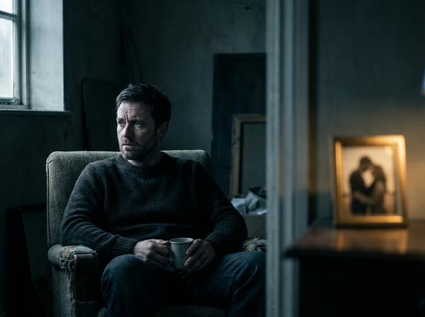 En mann i 50-årene som sitter alene i en halvmørk stue, med et ettertenksomt og distansert uttrykk, mens et varmt lys i bakgrunnen antyder et intimt par som står i kontrast til hans følelse av ensomhet.