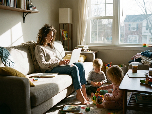 O mamă epuizată, cu un laptop în brațe, înconjurată de copiii săi mici care se joacă într-un living cald, dar haotic, simbolizând presiunea constantă și dorința de echilibru.